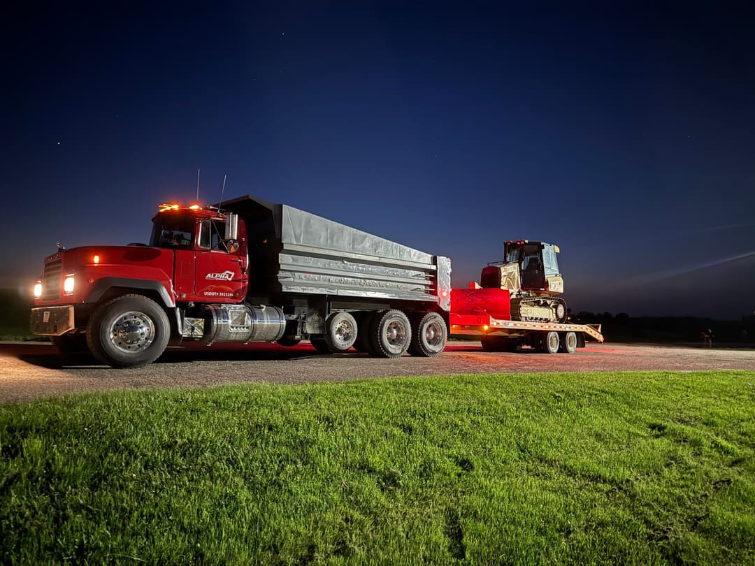 Red dump truck with a construction vehicle on a trailer parked on a roadside at night.