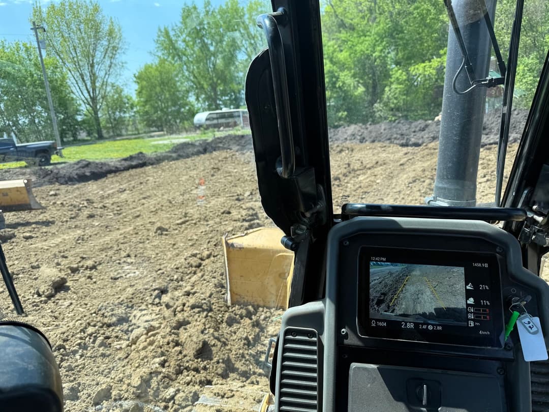 View from a construction vehicle's cabin showing a dirt excavation site and dashboard display.