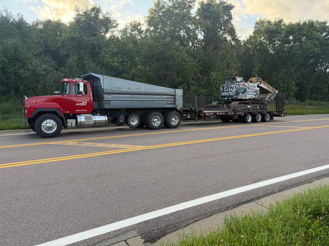 Red dump truck towing a flatbed with construction equipment on a roadside.
