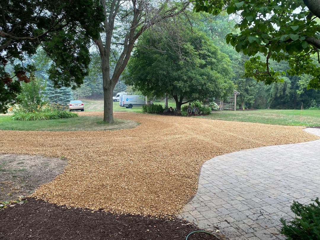 Gravel driveway installation in a landscaped yard with trees and a van in the background.