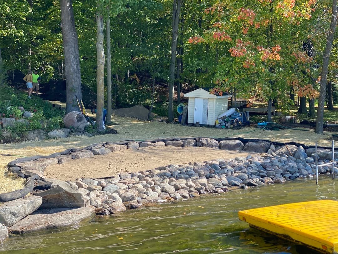 Scenic lakeshore with rocks, sand area, colorful trees, and a storage shed in background.